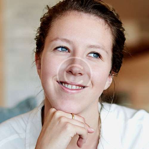 A woman with light brown hair smiles thoughtfully, resting her chin on her hand. She wears a white shirt, and the background is softly blurred, conveying a warm tone.