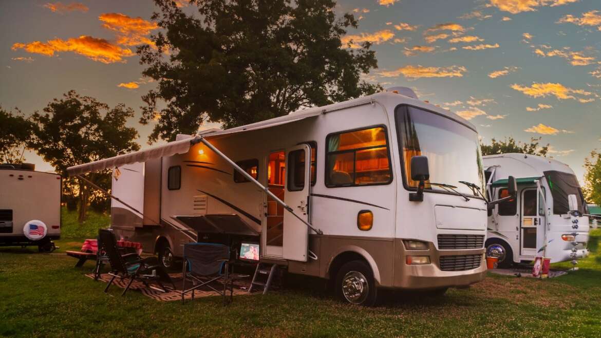 White RV parked on grass with awning and chairs, sunset sky in background.