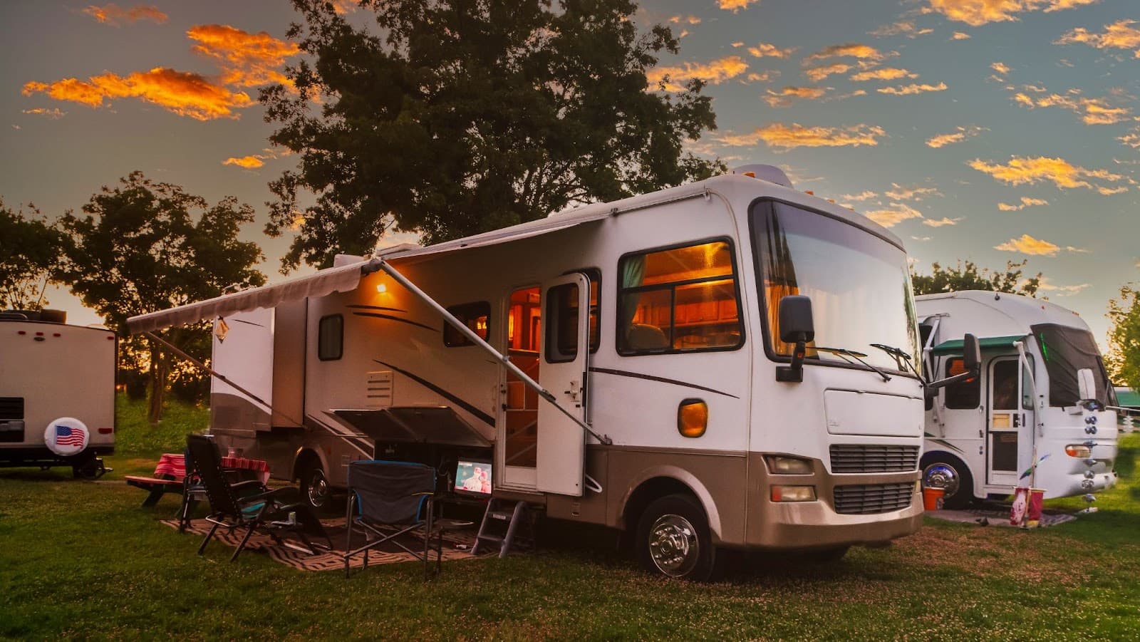 White RV parked on grass with awning and chairs, sunset sky in background.
