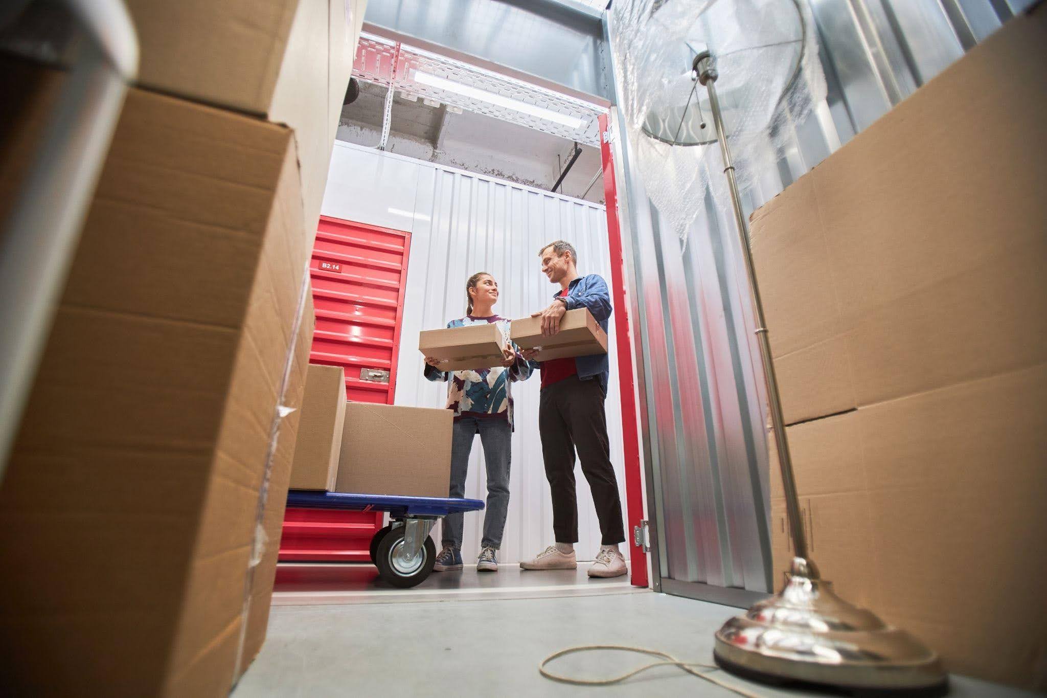 A man and woman are lifting boxes together inside a storage unit organizing their belongings