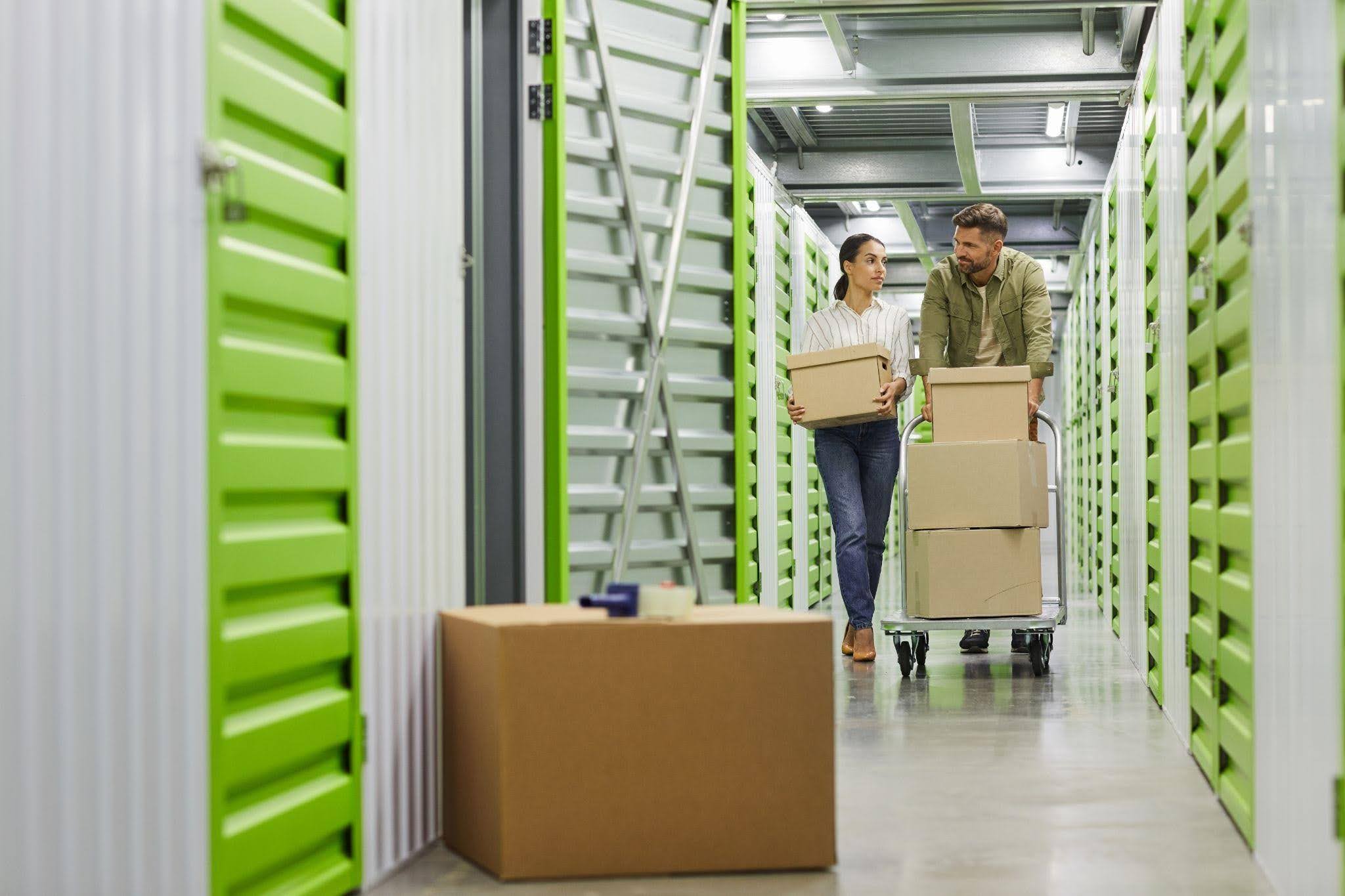 A man and woman navigate a storage unit filled with stacked boxes and miscellaneous items