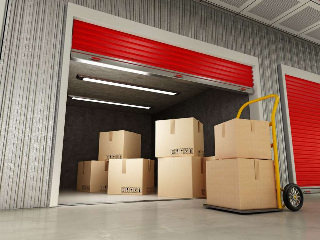 Open red storage unit door with stacked cardboard boxes and a yellow hand truck inside.